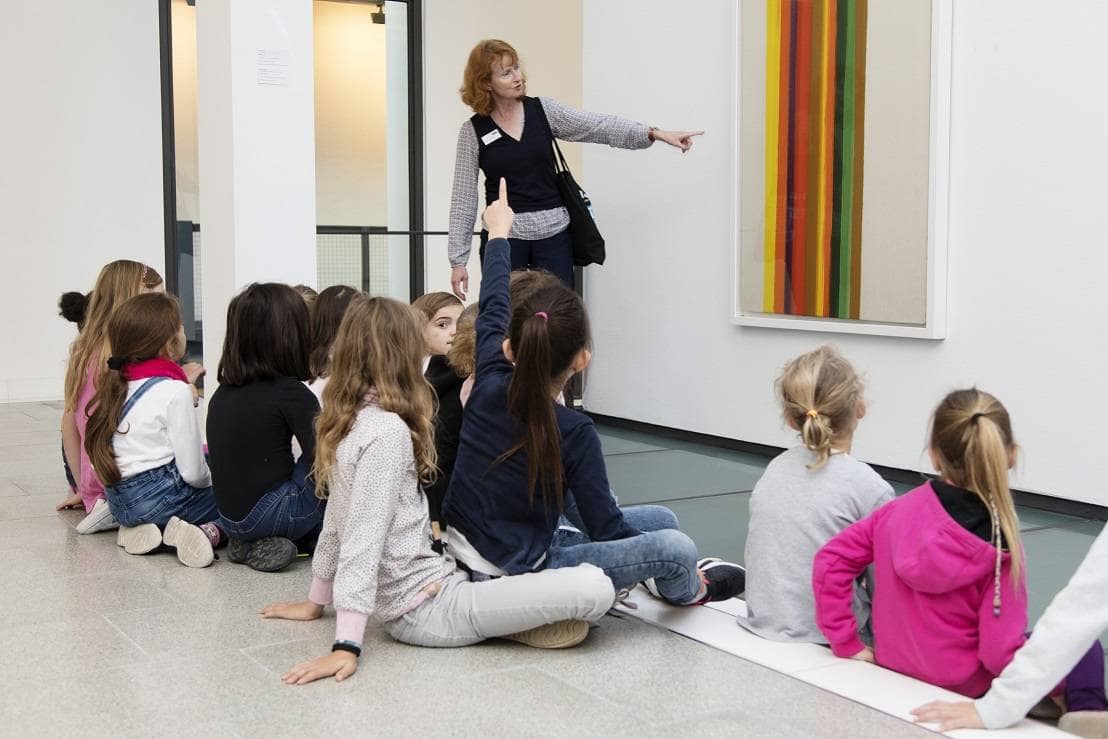 Kinder sitzen auf dem Boden vor einem Bild im Museum und bekommen von einer Theaterpädagogin etwas dazu erzählt